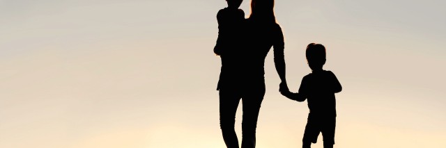 Silhouette of a young mother walking and lovingly holding hands with her happy little child, while holding his baby brother, outside in front of a sunset in the sky.