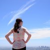 Half length portrait of young woman from behind. Woman is wearing sunglasses and a sleeveless shirt. Subject has her hands on her hips and is looking at a city skyline. Image is landscape orientation.