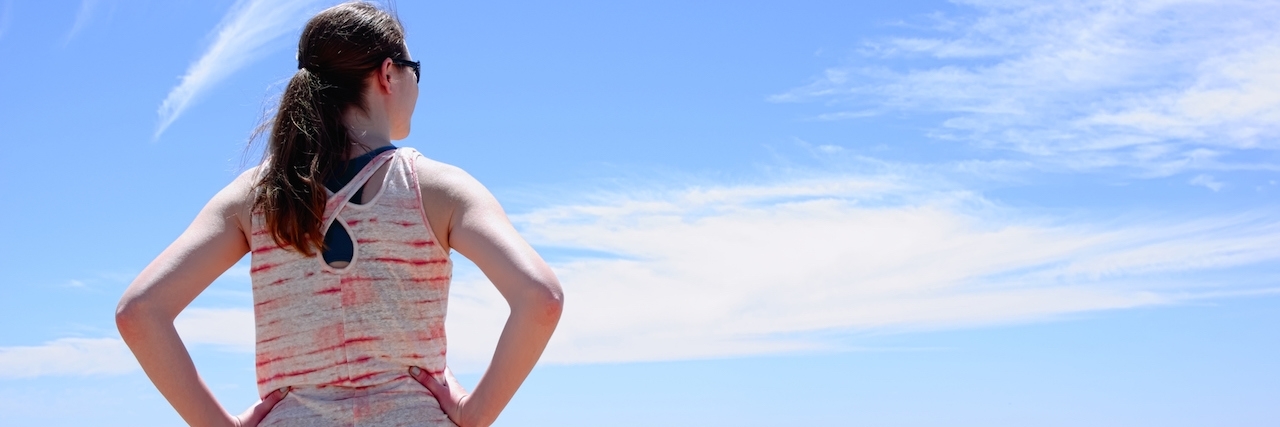 Half length portrait of young woman from behind. Woman is wearing sunglasses and a sleeveless shirt. Subject has her hands on her hips and is looking at a city skyline. Image is landscape orientation.