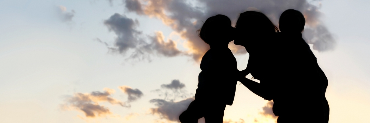 the Silhouette of a mother and her two young children; a little boy and his baby brother are playing outside at sunset, hugging and kissing.