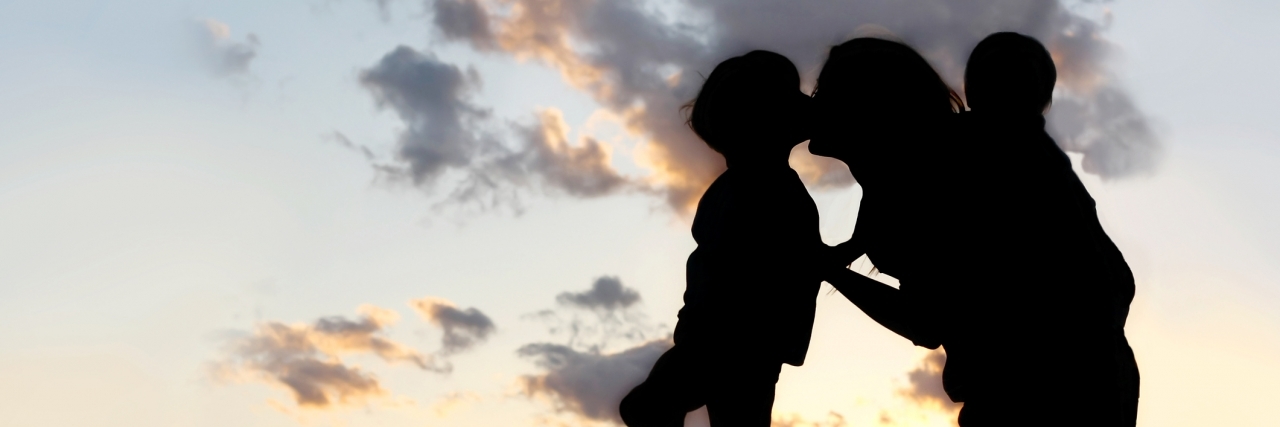 Letter to My Kids About Life With Lyme Disease and Lupus the Silhouette of a mother and her two young children; a little boy and his baby brother are playing outside at sunset, hugging and kissing.