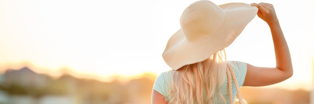 Why I'm Not Showing My Scars and Medical Devices in Public A slender woman,with long blonde hair in a straw hat with large brim,blue shorts and blue t-shirt,on the shoulder woven straw bag,admiring the sunset while standing on the beach near the sea