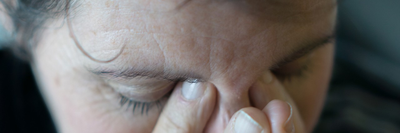 Losing a Child With PMM2-CDG, the Same Disorder as My Son A dark image and close up of a middle aged woman rubbing her eyes and holding her head, as if in anguish, or with a bad headache.