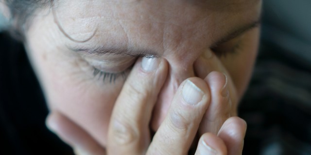 A dark image and close up of a middle aged woman rubbing her eyes and holding her head, as if in anguish, or with a bad headache.