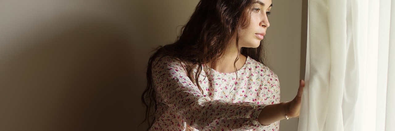 Questions to Ask Friend With Chronic Fatigue Syndrome Girl sitting alone on table near big window and looking outside thru white curtain