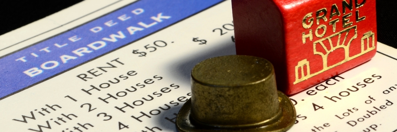 How Chronic Diseases Are Like Monopoly Game Detail of red wooden monopoly hotel and houses with top hat game piece and deed to boardwalk on black background