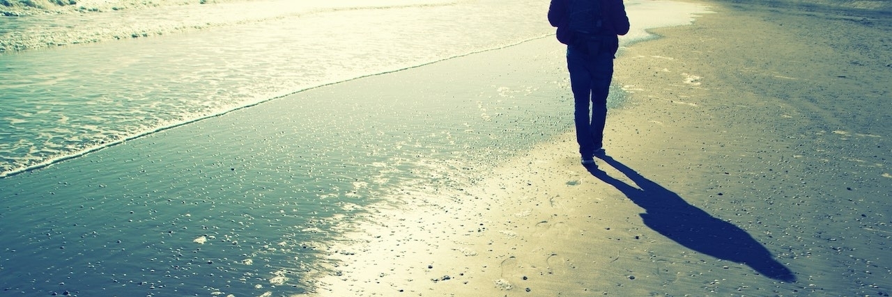 Guilt About Fibromyalgia Because It's Not Life-Threatening Person walking alone on sunny sandy beach with shells, foot prints and sea waves. Lonely walking on sea beach with waves at sunny day.