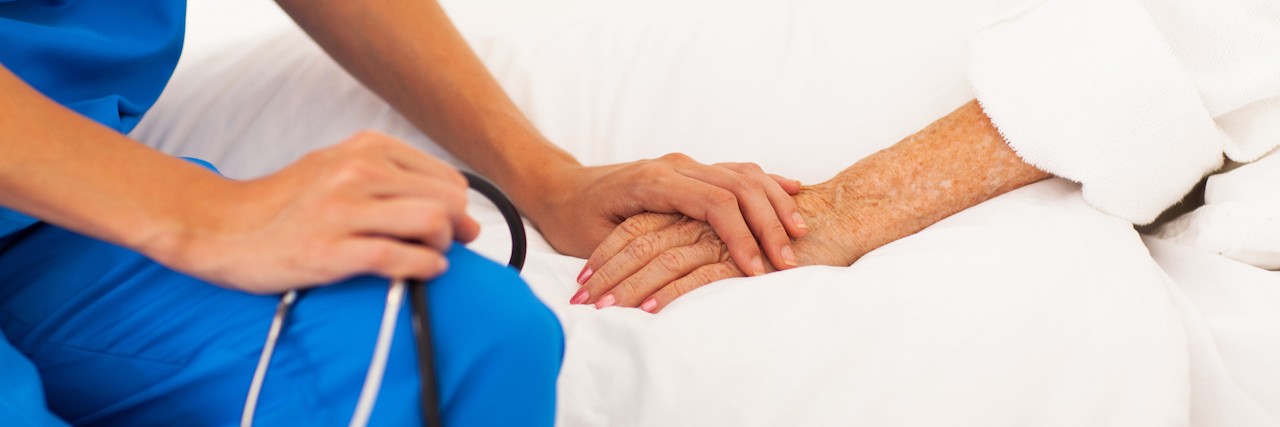 Nurse with stethoscope is show embracing the hand of a patient as they sit on the patient's bed