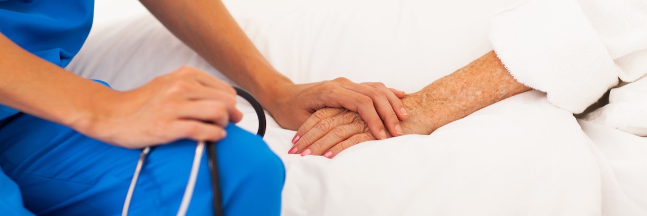 How My Scars Can Be Used to My Advantage as a Nurse Nurse with stethoscope is show embracing the hand of a patient as they sit on the patient's bed