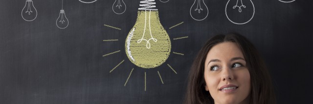 Young business woman standing with her back against a blackboard. She is looking to a big glowing light bulb.