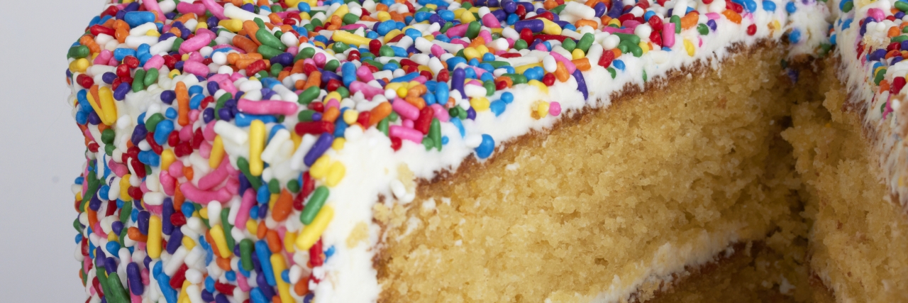 A yellow cake with vanilla frosting that is encrusted in rainbow sprinkles. The cake was shot against a pale gray backdrop.