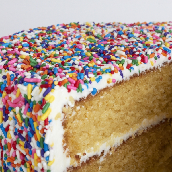 A yellow cake with vanilla frosting that is encrusted in rainbow sprinkles. The cake was shot against a pale gray backdrop.