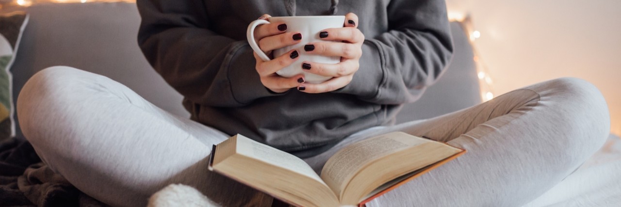 Fun Things to Do While Coping With Illness Girl holding cup of hot tea and reading in bed. Around her in bad earphones, book, smart phone. Decorative lights in background.