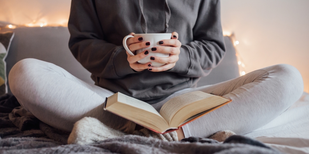 Girl holding cup of hot tea and reading in bed. Around her in bad earphones, book, smart phone. Decorative lights in background.