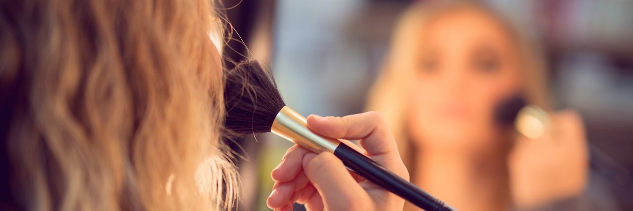 Why Wearing Makeup Makes Me Feel Better About My Fibromyalgia woman putting makeup on in front of mirror