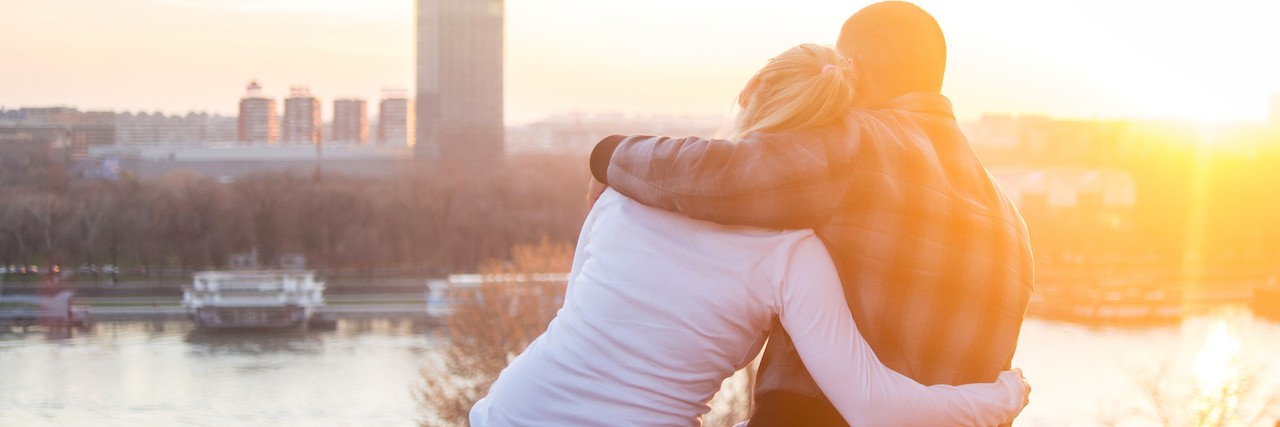 How to Help Friend or Family Cope With a New Diagnosis Rear view of a loving couple sitting on a ledge and looking at river view from above.