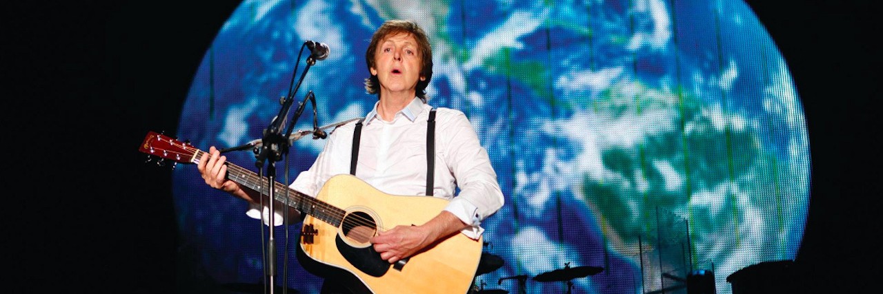 Paul McCartney holding a guitar, standing in front of a photo of the world