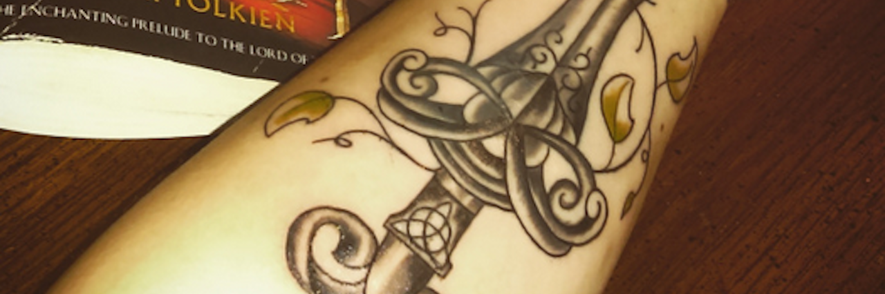 When I Wasn't Ready to Get a Semicolon Tattoo Woman shows her sword tattoo on her forearm, with book and coffee next to her