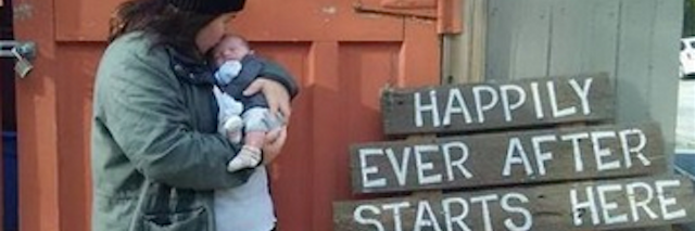 Woman holding baby in front of wooden door and sign that says "Happily ever after starts here."