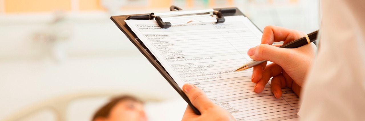 What I Wish the Doctors in the Hospital Understood About Lyme Disease Close up of doctor writing on a medical chart with patient lying in a hospital bed in the background