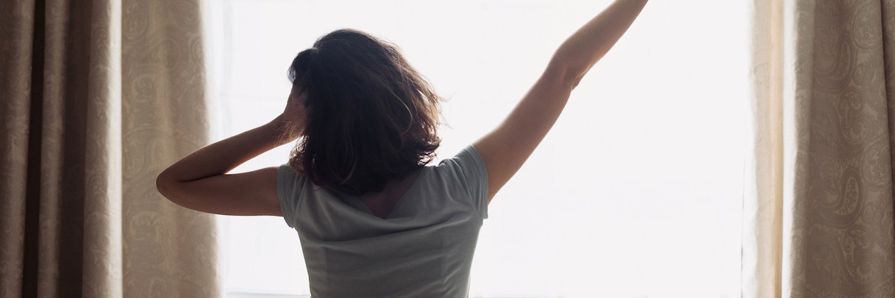 When You Feel Better in the Morning With Chronic Illness Young woman waking up and stretching on her bed, back view
