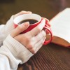 A close-up of hands holding a red mug filled with coffee on a wooden table next to an open book