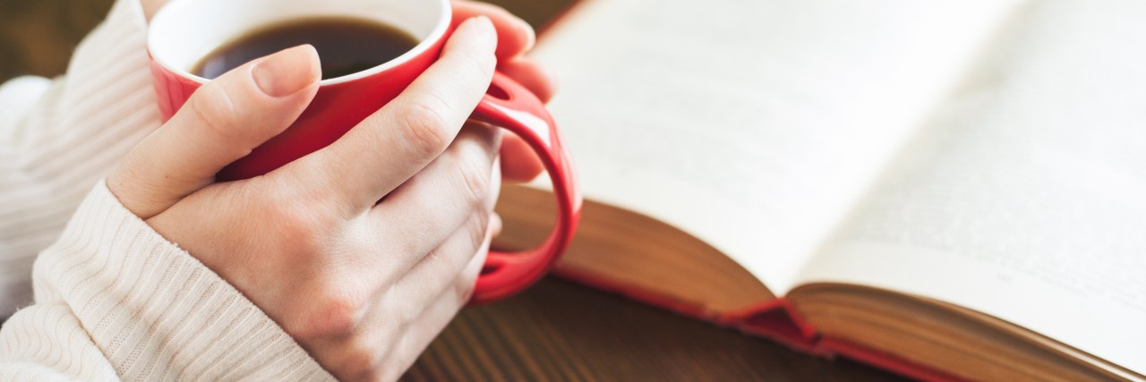 Ways to Take Care of Yourself as a Caregiver A close-up of hands holding a red mug filled with coffee on a wooden table next to an open book