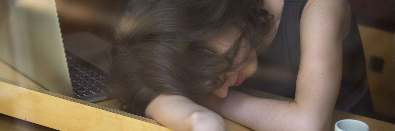 Common Mistakes About Pacing Yourself When You're Sick Young woman lying on her arms on the table in cafe in front of laptop with cup of coffee, sleepy, tired, overworked