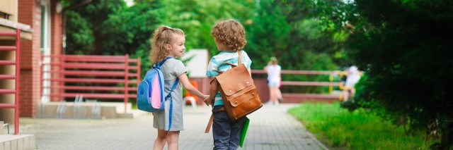 Young students, boy and girl, going to school. They hold hands. They hold hands. Children behind shoulders have satchels. Warm day in an early autumn. Back to school.
