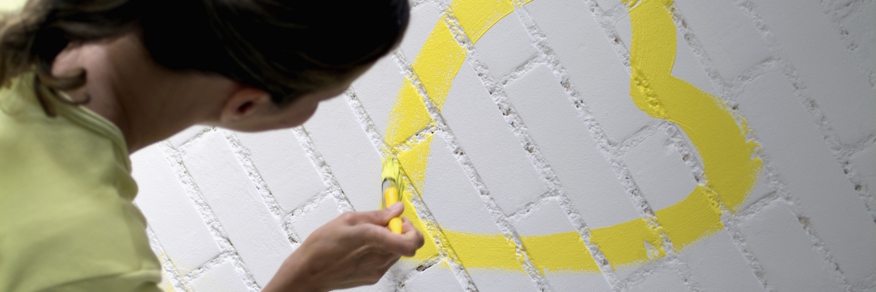 Rear view of a woman painting a yellow heart on a brick wall