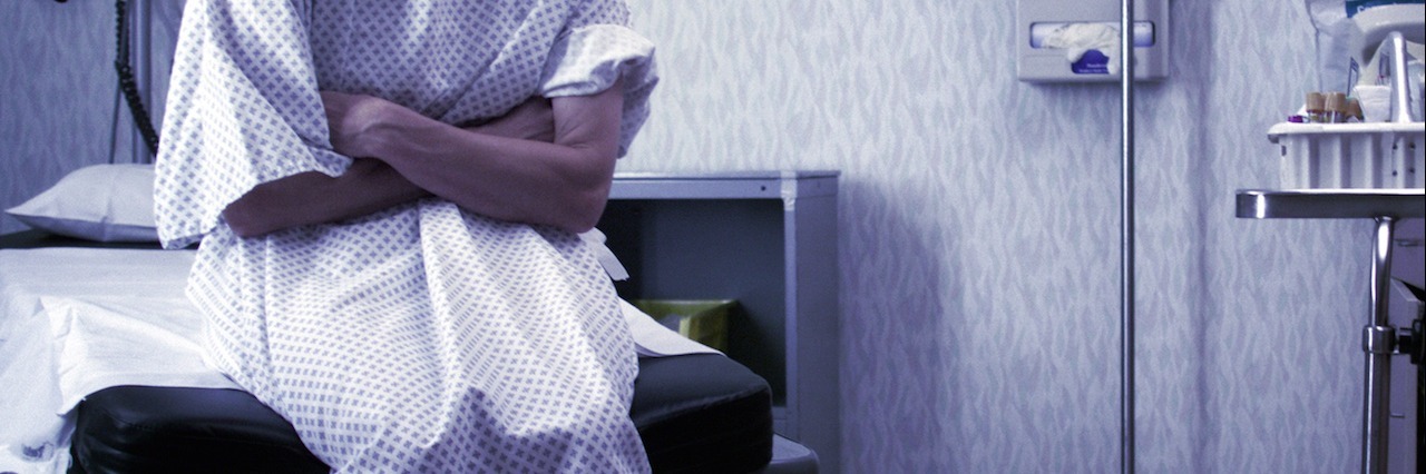 What It's Like Going to the Emergency Room With a Chronic Illness woman sitting on exam table at hospital wearing hospital gown