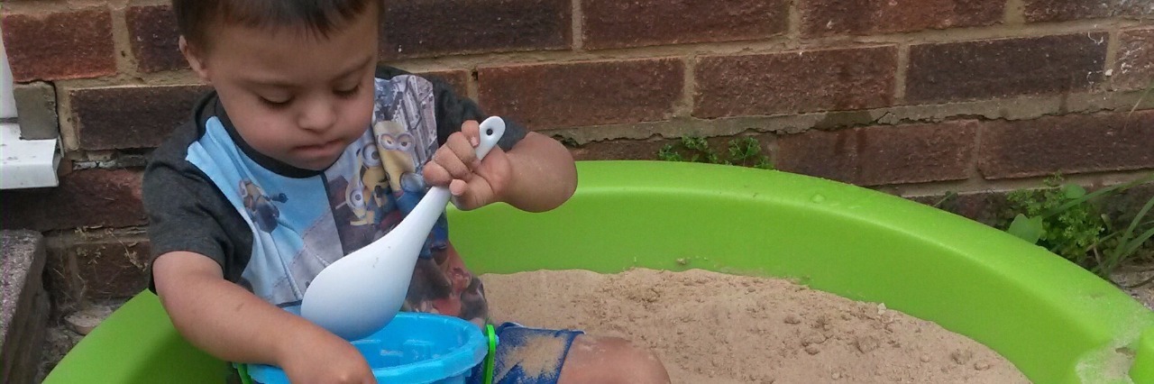 The Summer Before My Son With Down Syndrome Starts School Boy sitting in a sand pit playing with a spoon and bucket.