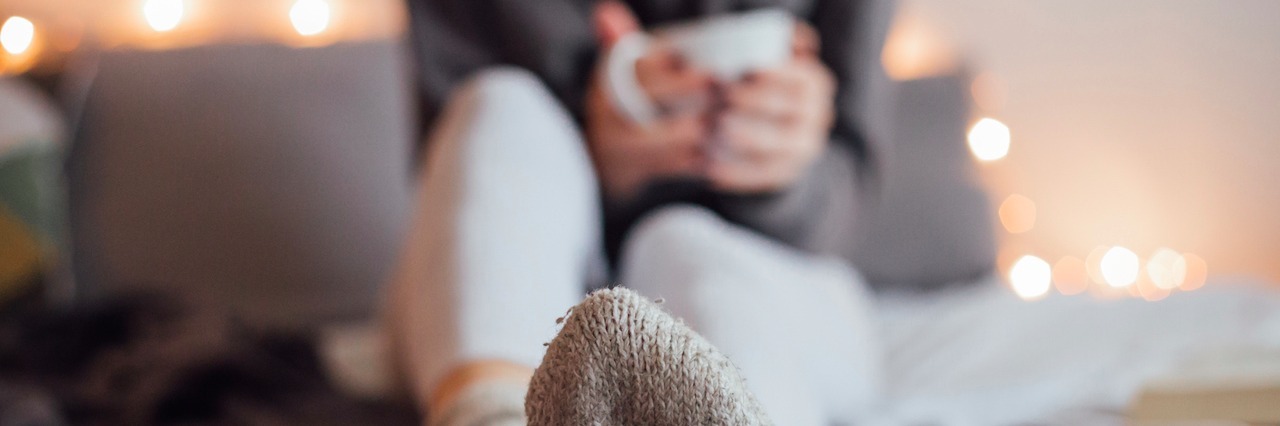 Little Things That Help Me Cope With Being Sick Girl holding cup of hot tea and reading in bad. Around her in bed book, smart phone. Decorative lights in background.