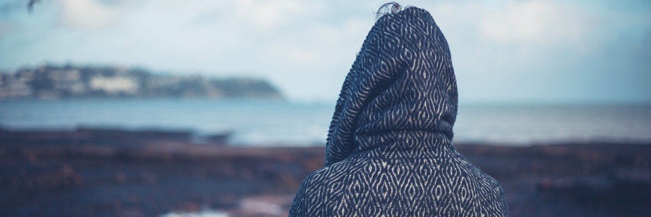 How to Cope With Depression and Chronic Illness A young woman wearing a hooded coat is walking on a dramatic beach in the winter