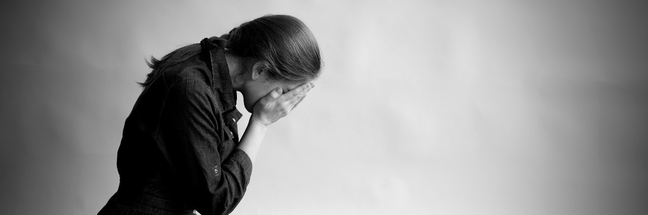 Woman is sitting in profile on an old cracky floor. She is sad and depressed, crying and covering her face with hands. Studio paper background in behing her.
