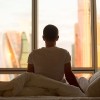 Rear view of young man sitting on unmade bed and looking at dawn city scenery in window after waking up.