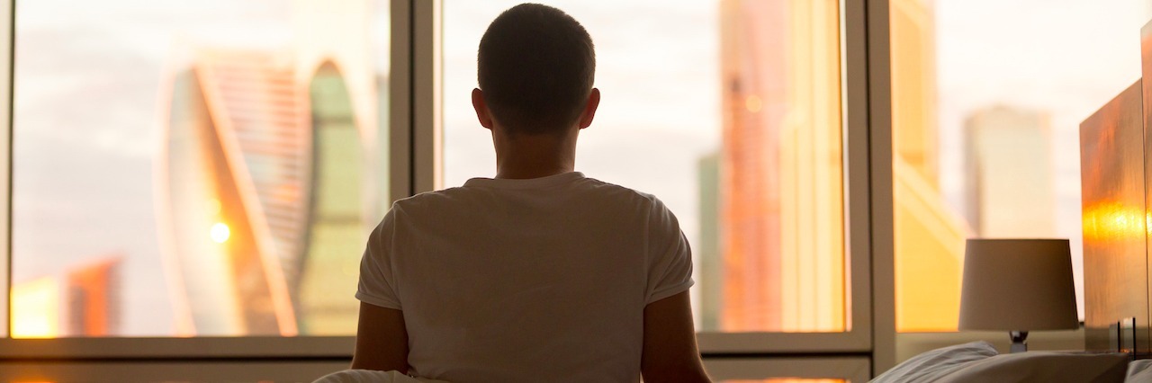 Rear view of young man sitting on unmade bed and looking at dawn city scenery in window after waking up.