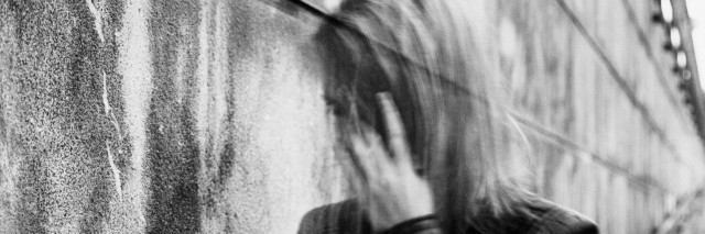 Young woman experiencing a severe disorientation, confusion, or sadness outdoors, in front of a wall. Converted to black and white, grain added, blurry, slightly out of focus.