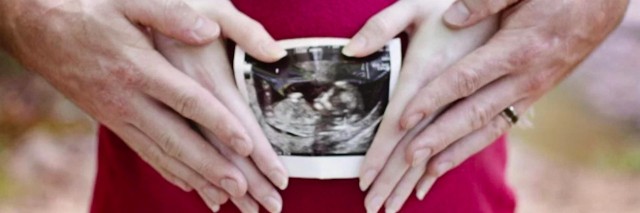 Two pairs of hands on a pregnant woman's stomach, holding an ultrasound photo