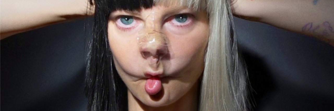 How Sia's Music Helps Me in Eating Disorder Recovery A photo of a young woman with tape on her nose and her hands on top of the half black, half white wig on her head