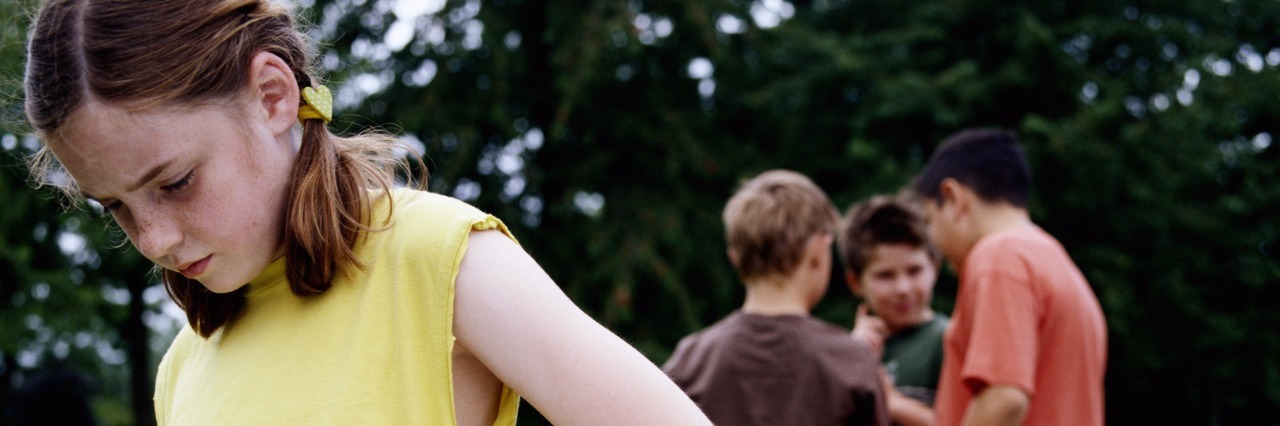 Girl (9-11) in park hand on hip, looking down, boys in background