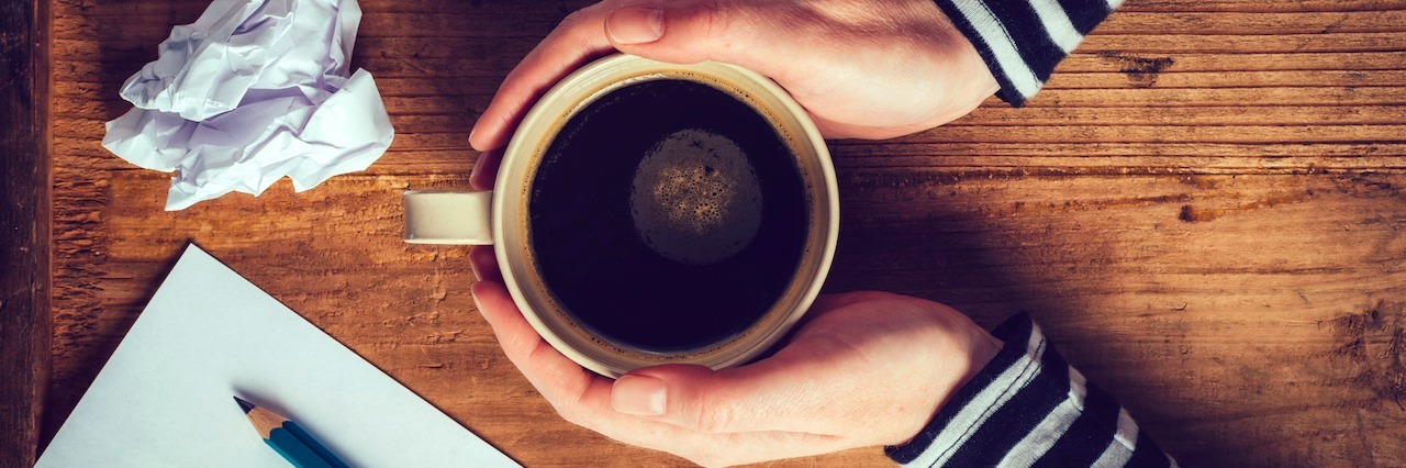 A Letter to My Anxiety About the Reasons It Is Wrong woman holding a cut of coffee with a pen and crumpled up pieces of paper on her desk