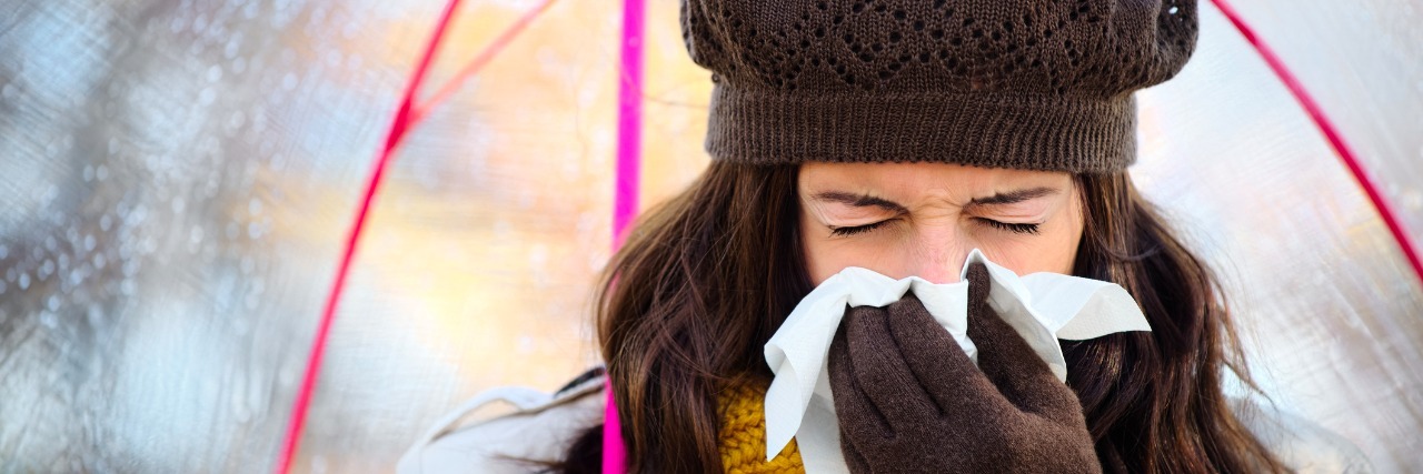 Feeling Anxiety in Cold and Flu Season Woman with cold or flu coughing and blowing her nose with a tissue under autumn rain. Brunette female sneezing and wearing warm clothes.