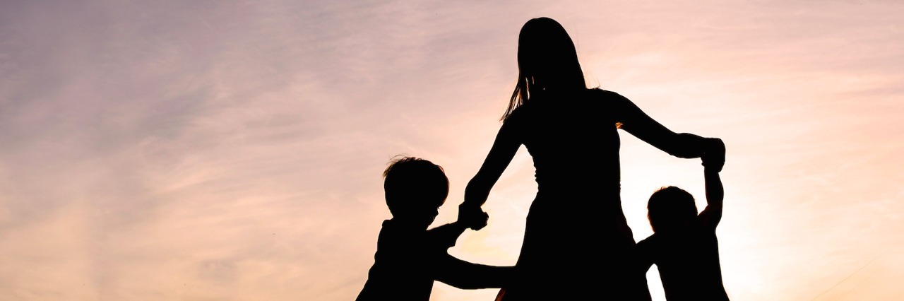 Aunt Asks How Loved Ones Can Help Support Autistic Children Silhouette of a woman and two young children holding hands and dancing around outside, isolated against the sunset.