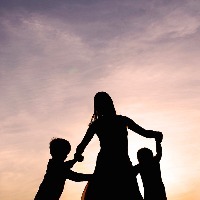 Silhouette of a woman and two young children holding hands and dancing around outside, isolated against the sunset.