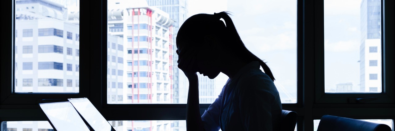 When Co-Workers Witnessed My Panic Attack side profile of a woman sitting at her desk in an office with her head in her hands