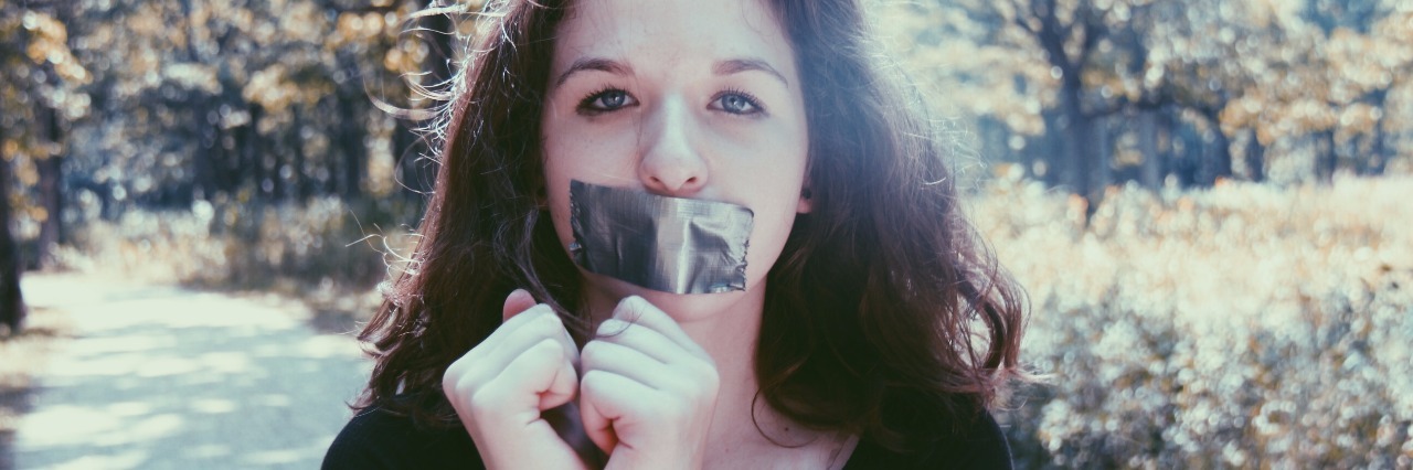 Losing Your Old Identity After Being Diagnosed With a Disease Kaitlyn standing outside in front of trees with duct tape over her mouth and binding her hands together