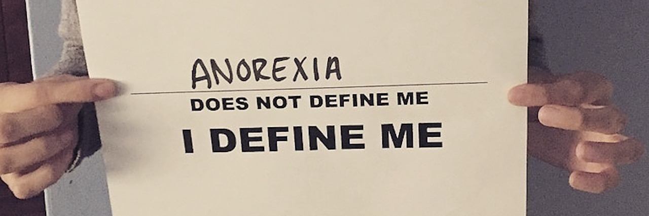 An upclose photo of a woman holding a sign that says, "Anorexia does not define me. I define me."