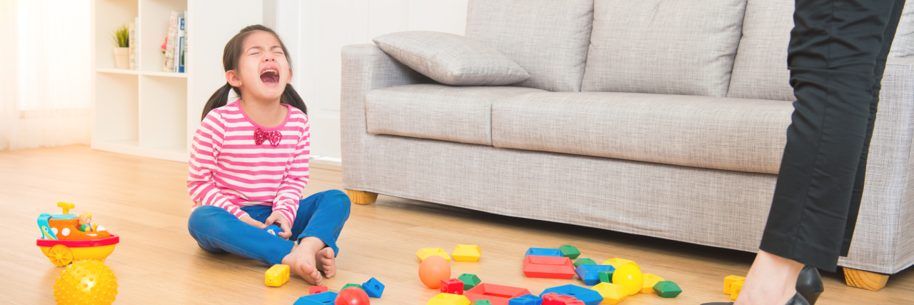 Girl angry and crying on the floor, surrounded by blocks, side of mom wearing a suit and pointing at her child