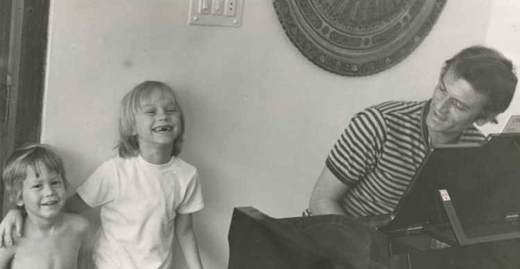 Two small kids standing next to their dad, who is seated at a piano, as they all laugh